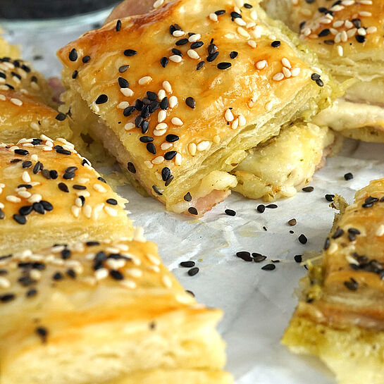 Leckerer Blätterteigsnack mit Käse, zartem Schinken und würzigem Pesto, mit goldbrauner Kruste und Sesam.