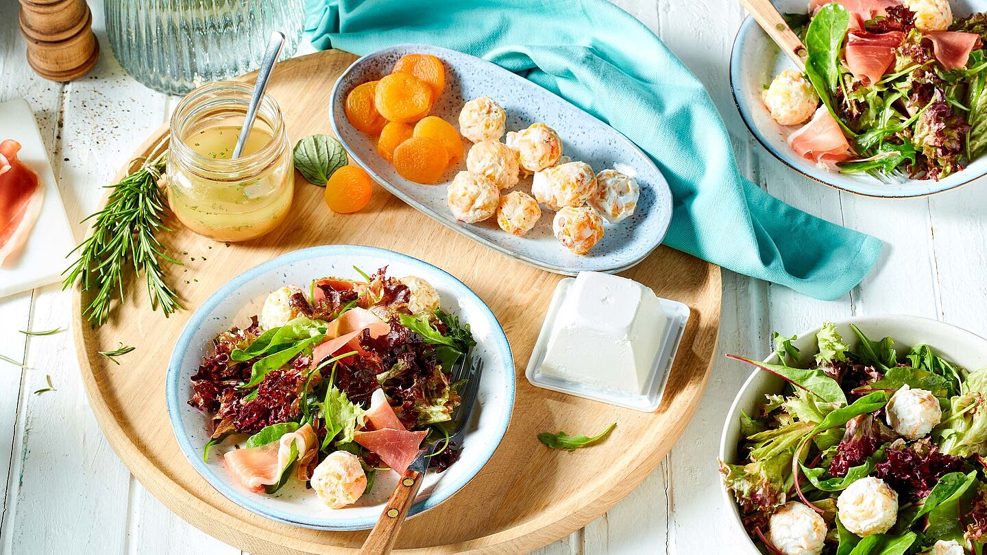 Holztablett mit einem leichten Sommersalat, der mit Aprikosen-Frischkäse-Bällchen und Serrano-Schinken garniert ist.