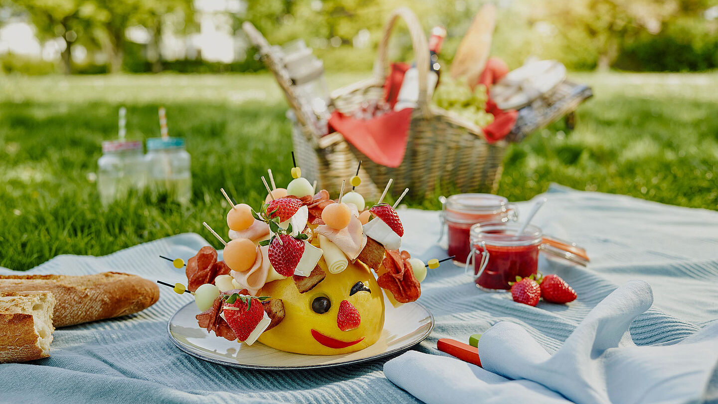 Käseigel Gérard hat auf seinen Spießen Géramont, Honigmelone und Putenbrust und sitzt auf einer Picknickdecke