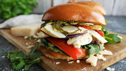 Mediterraner Burger mit gegrilltem Hühnchen, Gemüse, Rucola, Parmesan und Pesto auf einem Holzbrett angerichtet.