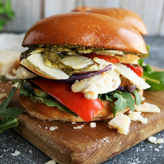 Mediterraner Burger mit gegrilltem Hühnchen, Gemüse, Rucola, Parmesan und Pesto auf einem Holzbrett angerichtet.