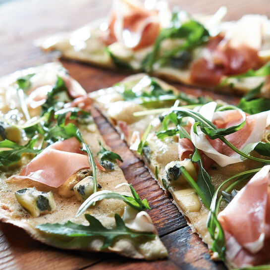 Dünner Flammkuchen mit Rucola und Bauernschinken auf einer rustikalen Holzunterlage 