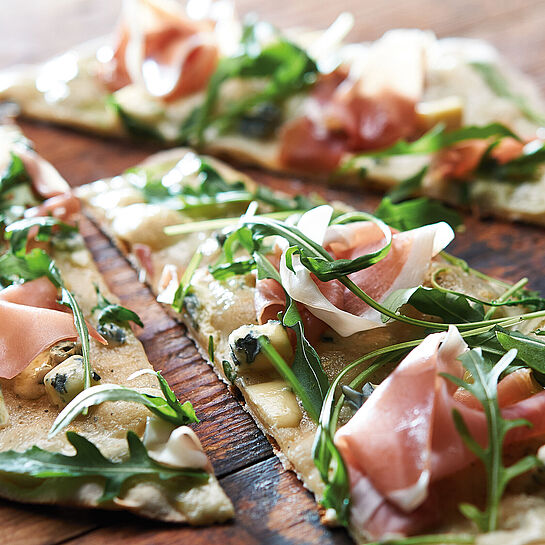 Dünner Flammkuchen mit Rucola und Bauernschinken auf einer rustikalen Holzunterlage 