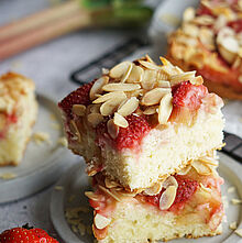 Ein Stück Rhabarber Blechkuchen mit Erdbeeren und knusprigen Mandelblättchen, bestreut mit Puderzucker, liegt auf einem Kuchengitter.