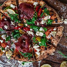 Pizza belegt mit hauchdünnen Bresaola Schinken Scheiben, frischem Rucola, Kirschtomaten, Zitronenscheiben und Peyrigoux Käse.