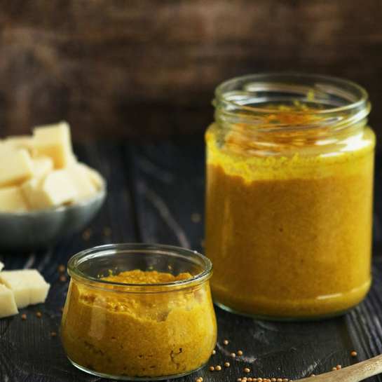 Zwei Gläser mit selbstgemachtem scharfem Senf, daneben ein Holzlöffel mit Senfkörnern und eine Schale mit Käsewürfeln auf dunklem Holzuntergrund.