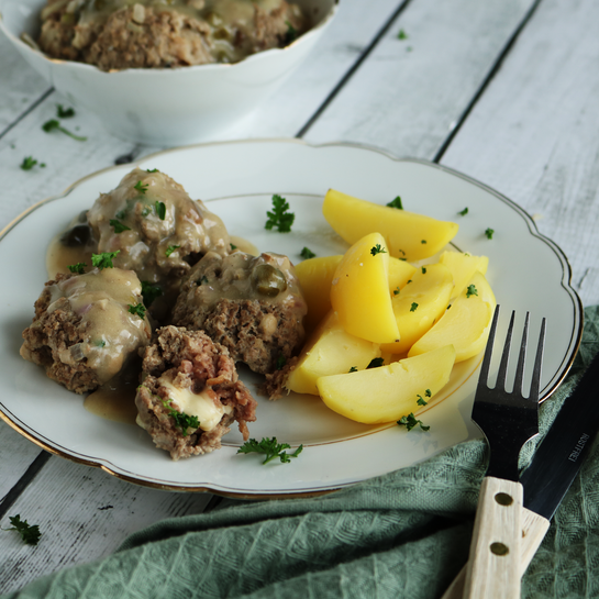 Teller mit Goldrand drauf liegen Königsberger Klopse mit Kapernsoße und Salzkartoffelspalten mit Petersilie. 