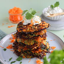 Knusprige Zucchini-Möhren-Puffer, garniert mit einem Klecks Kräuterfrischkäse und frischem Koriander, auf einem weißen Teller serviert.​