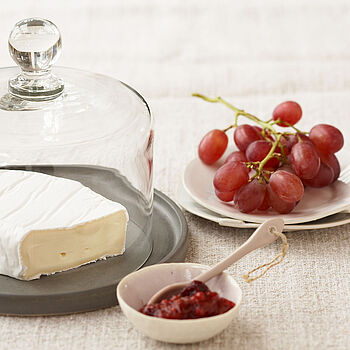 Unter einer Käseglocke aus Glas wird ein Camembert Käse serviert, dazu Weintrauben und Marmelade