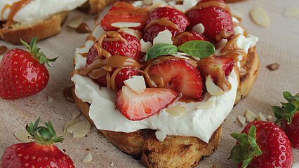 Knuspriges Brioche mit cremiger Frischkäsecreme, marinierten Balsamico-Erdbeeren, Salzkaramell und gehackten Mandeln, garniert mit Basilikum.