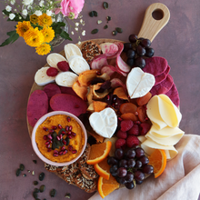 Kreatives Käseboard mit Brie, Trauben, Orangen, Himbeeren, Crackern und Dip auf Holzbrett, dekoriert mit Blumen.