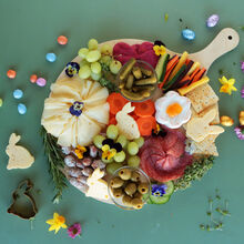 Runde Holzplatte mit einer dekorativen Oster-Snack-Auswahl, darunter Käse in Hasen- und Blumenform, Salami, Gemüse, Trauben, Oliven, Crackern und kleinen Deko-Ostereiern, arrangiert auf grünem Hintergrund und verziert mit essbaren Blüten.