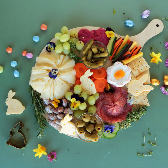 Runde Holzplatte mit einer dekorativen Oster-Snack-Auswahl, darunter Käse in Hasen- und Blumenform, Salami, Gemüse, Trauben, Oliven, Crackern und kleinen Deko-Ostereiern, arrangiert auf grünem Hintergrund und verziert mit essbaren Blüten.