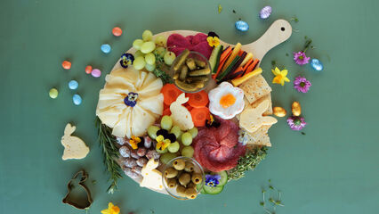 Runde Holzplatte mit einer dekorativen Oster-Snack-Auswahl, darunter Käse in Hasen- und Blumenform, Salami, Gemüse, Trauben, Oliven, Crackern und kleinen Deko-Ostereiern, arrangiert auf grünem Hintergrund und verziert mit essbaren Blüten.