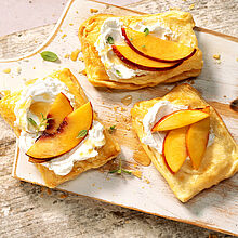 Süß-herzhafter Snack ► Blätterteig-Teilchen mit Ziegenkäse und Nektarinen, verfeinert mit Honig. Serviert auf einem Schneidebrett.